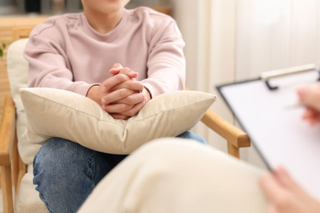 A person sitting with hands clasped on a pillow during a therapy session, facing a counselor holding a clipboard in a calm, supportive environment.
