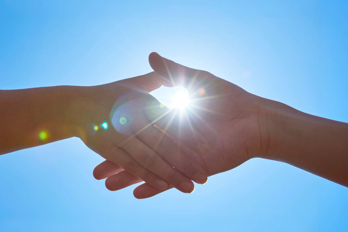 Two outstretched hands reach toward each other against a bright blue sky, with sunlight flaring brightly between them, symbolizing support and connection.