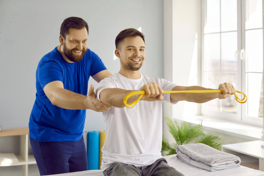 A smiling physical therapist in a blue shirt assists a young man in stretching a yellow resistance band during a rehabilitation exercise session in a bright clinical room.