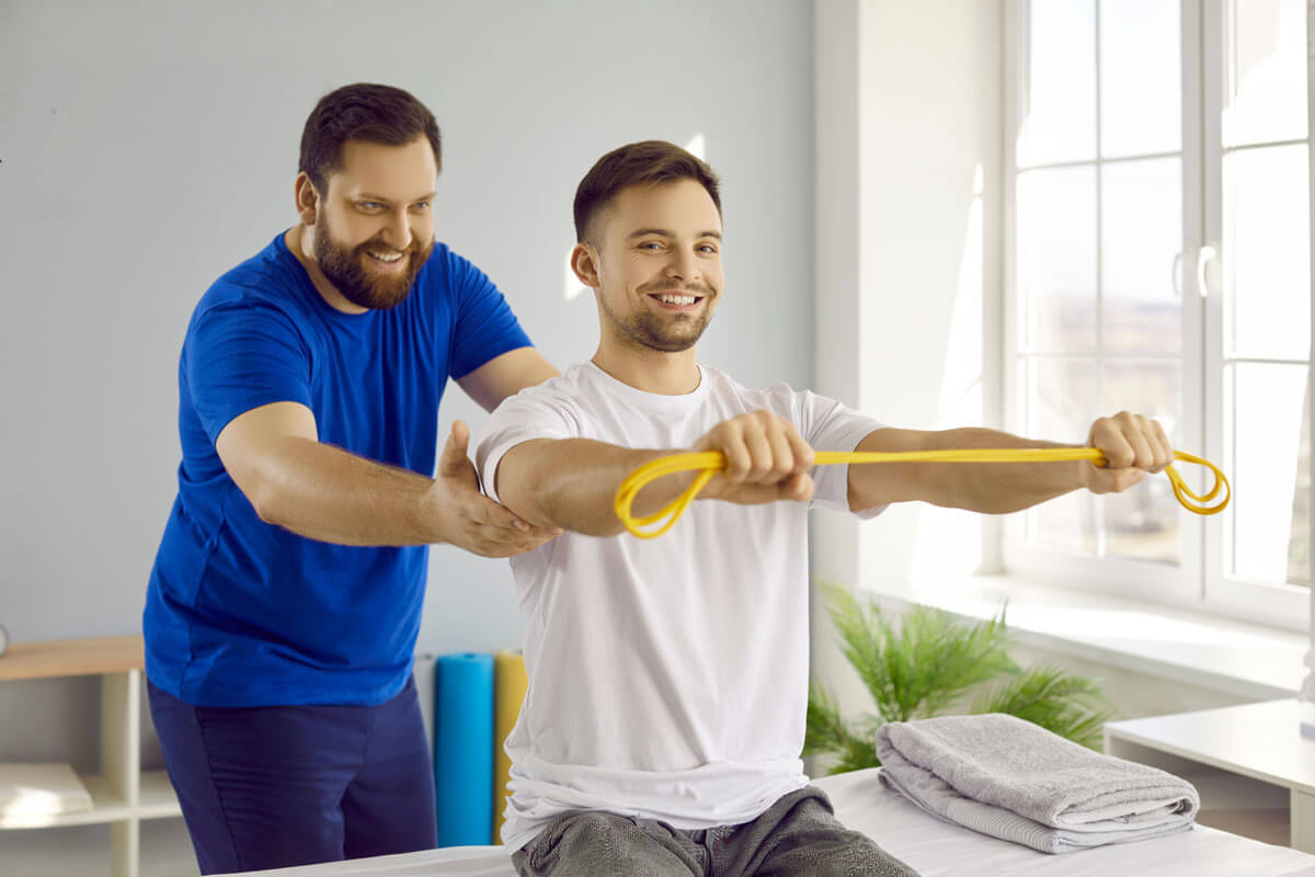 A smiling physical therapist in a blue shirt assists a young man in stretching a yellow resistance band during a rehabilitation exercise session in a bright clinical room.