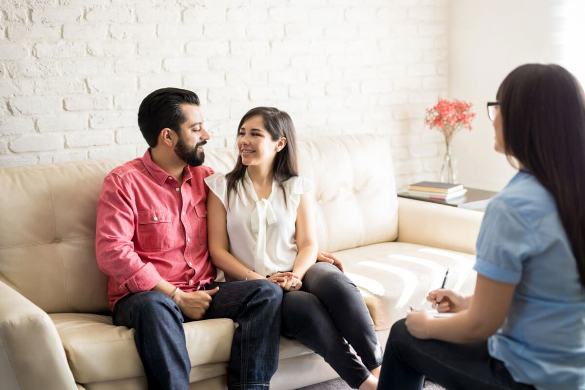 A smiling couple sitting close together on a couch during a counseling session with a therapist taking notes.