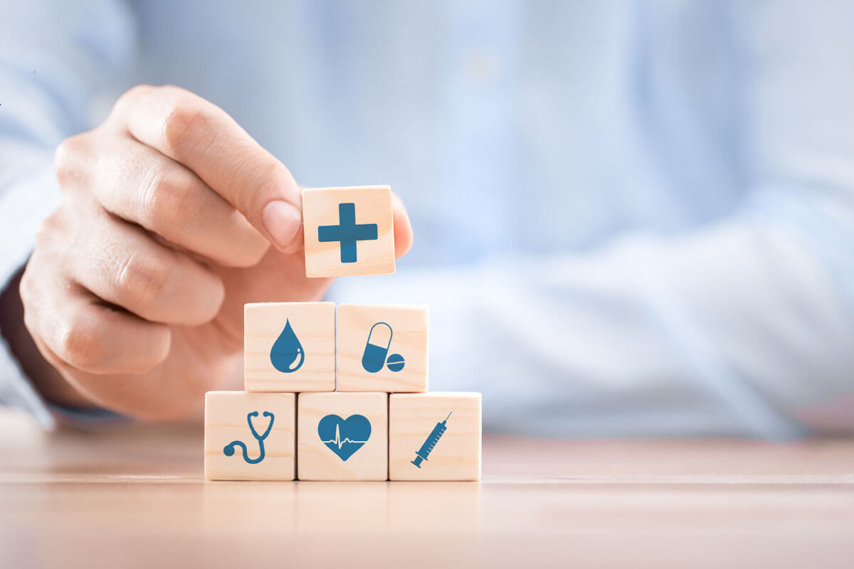 A hand stacks wooden blocks printed with medical icons like a heart, syringe, and stethoscope.