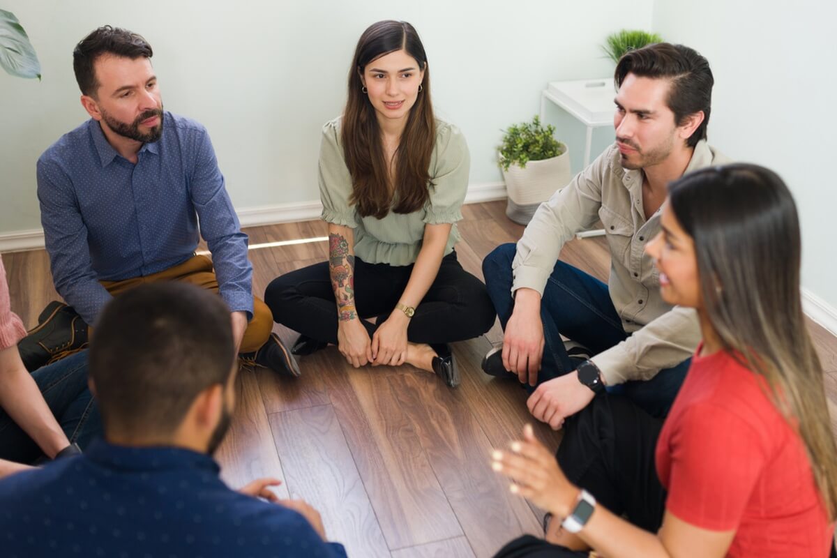 A group of people sit on a wood floor in a circle, listening during a group discussion.
